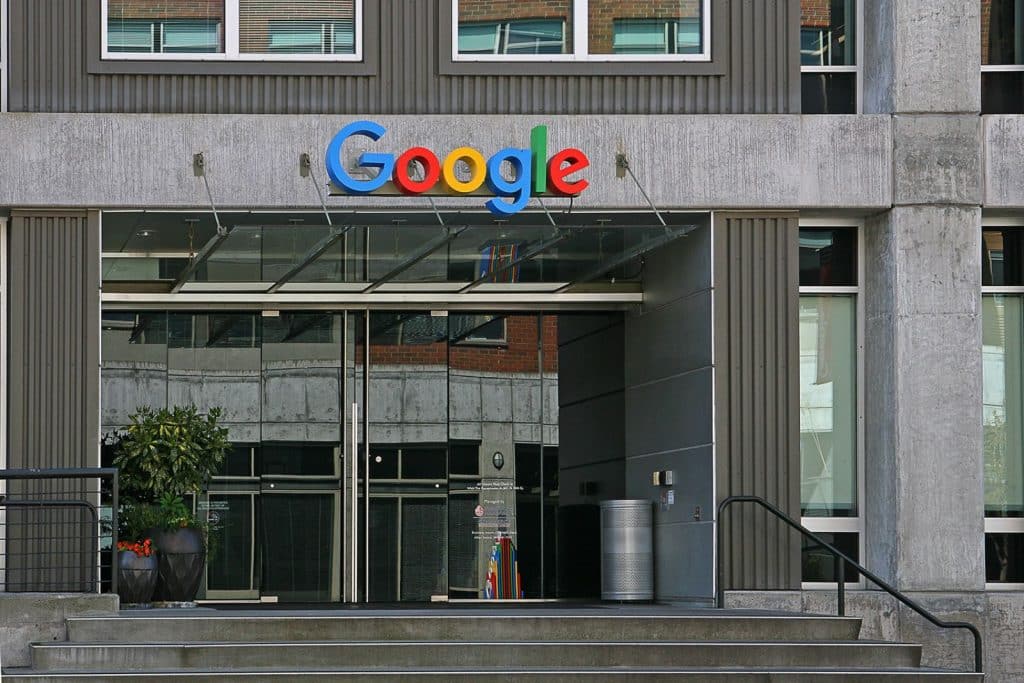 Entrance of a building featuring the Google logo above glass doors, with potted plants and steps leading up to the entrance.