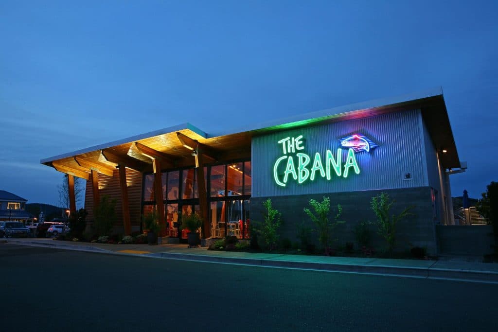 Exterior of "The Cabana" restaurant at dusk, featuring illuminated neon signage on a modern building with large glass windows and wooden accents.