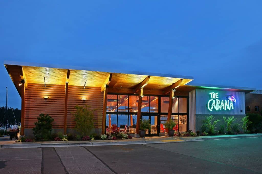 A modern building with wooden panels and large glass windows at dusk. A neon sign reads "The Cabana.