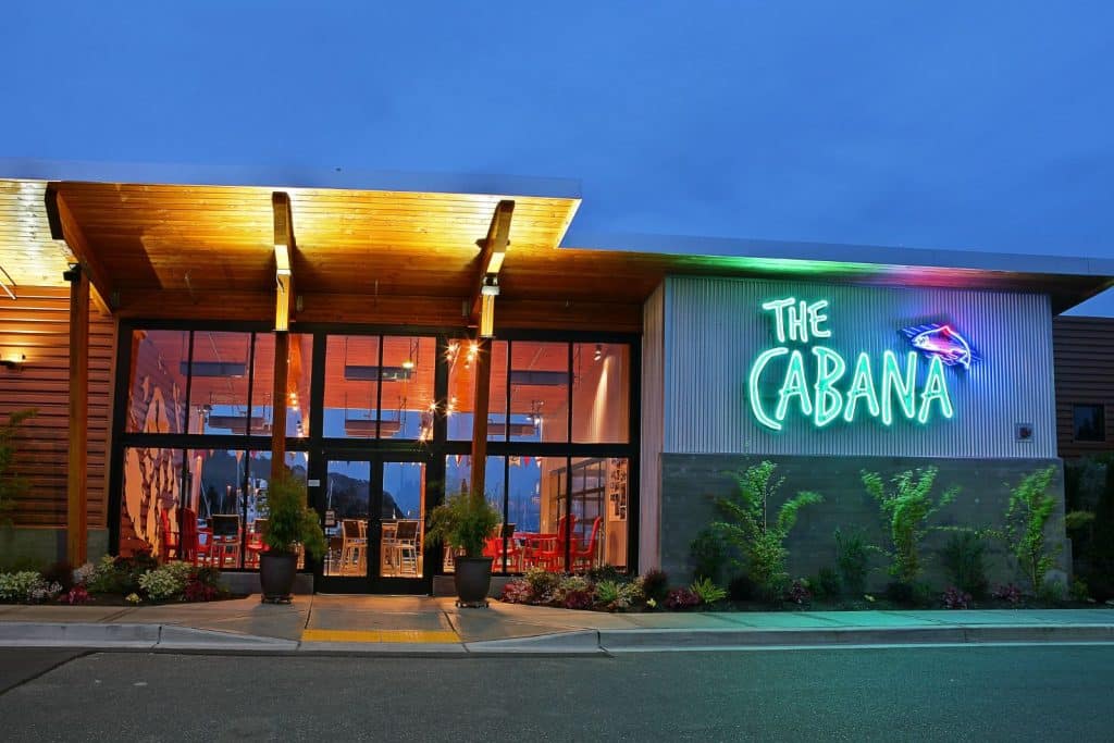 Exterior view of a modern building with large glass windows, featuring a neon sign that reads "The Cabana" against a blue evening sky.