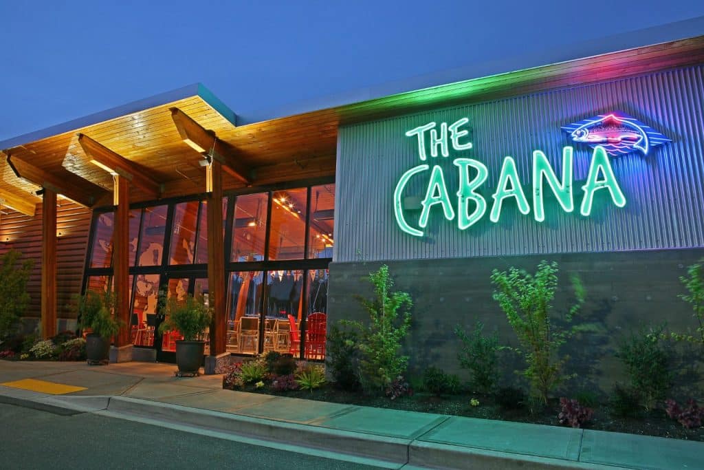 Exterior view of a building with a neon sign reading "The Cabana" at dusk. Large glass windows and plants adorn the entrance.