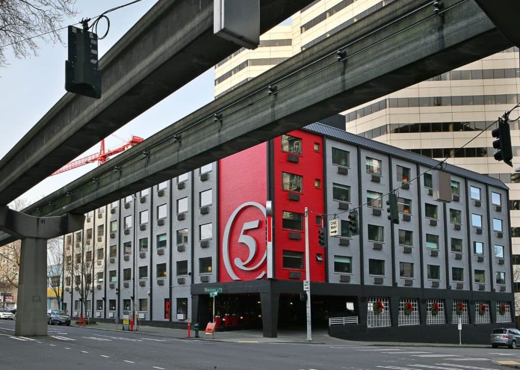 A modern building with a large number "5" on a red section of the facade is located under raised tracks and next to a busy urban street.