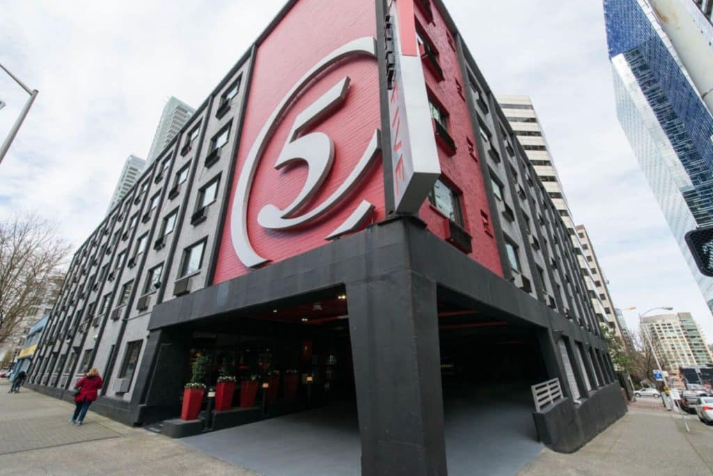 Exterior view of a building with a large red and gray "5" logo on the corner. The building has geometric patterns and is surrounded by city structures and pedestrians.