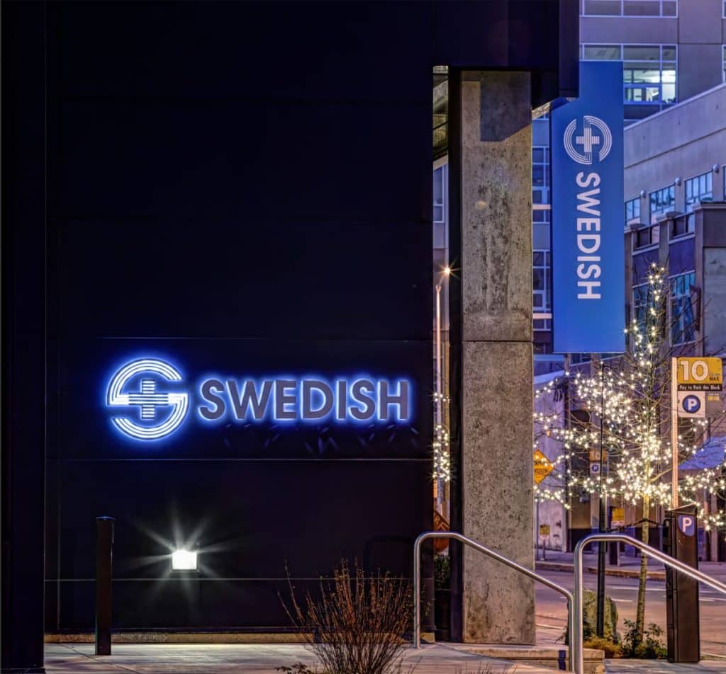 Illuminated "Swedish" sign on a dark building exterior with a blue vertical banner. Nearby trees are decorated with string lights.