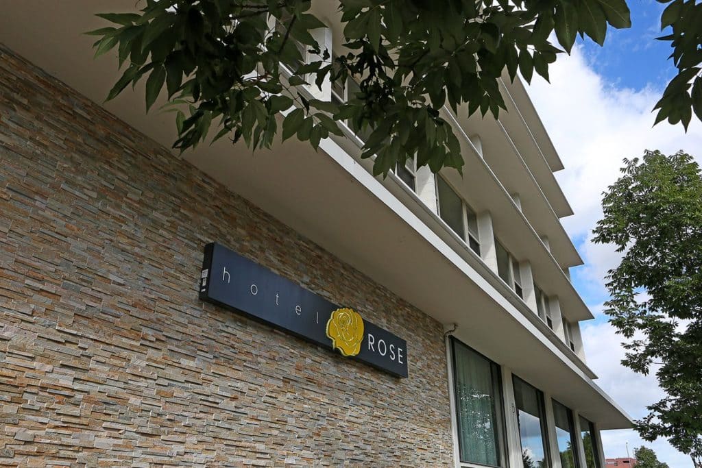 Exterior of a building with a stone facade and a sign that reads "hotel ROSE" featuring a yellow rose logo, surrounded by leafy trees.