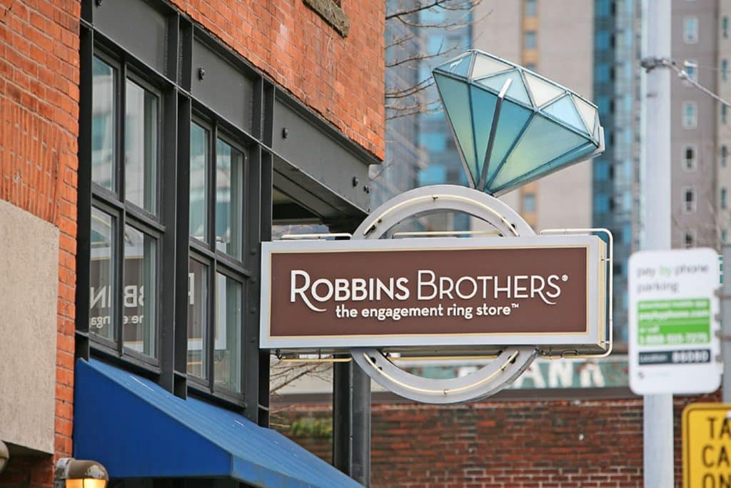 Sign for Robbins Brothers, the engagement ring store, mounted on a building with a large diamond decoration above it; city buildings and a parking meter in the background.