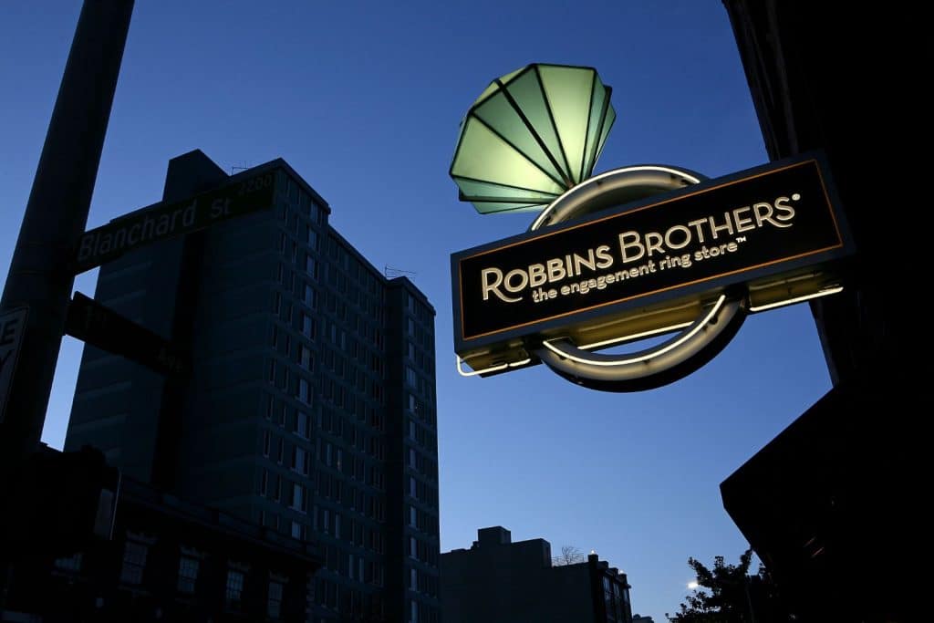 Illuminated sign for Robbins Brothers, the engagement ring store, against a backdrop of a city street with buildings at dusk.