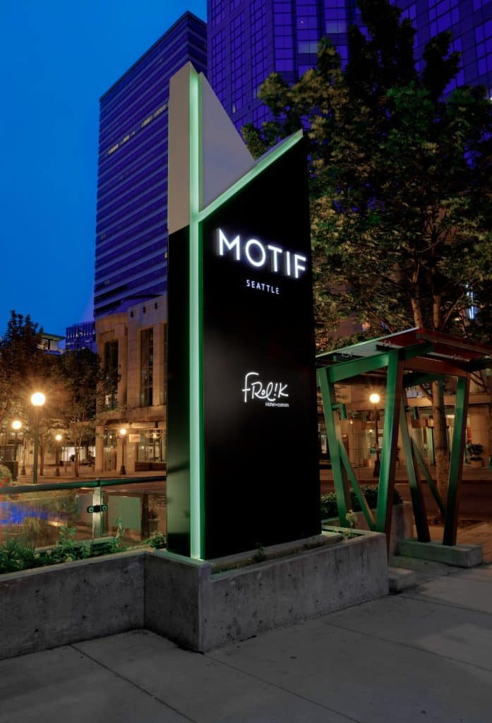 A black and green sign for Motif Seattle hotel with "Frolik Kitchen + Cocktails" below, set at an urban streetscape scene in the evening.