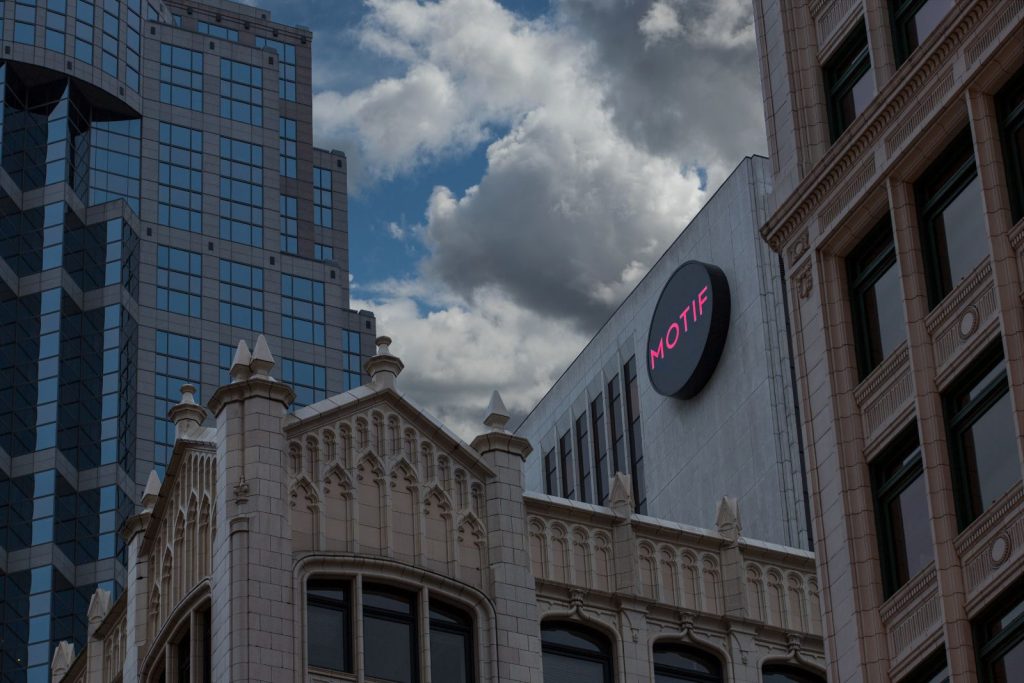 Urban skyline with three buildings; one features an ornate facade, another has reflective glass, and the third displays a sign reading "MOTIF" against a cloudy sky.