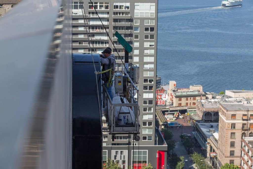 A worker stands on a suspended platform high above ground, attached to a building's facade, with a cityscape and body of water in the background.