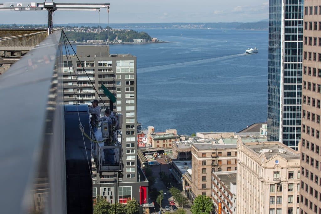 Workers on a suspended platform clean windows on a high-rise building overlooking a waterfront with boats and distant shoreline.