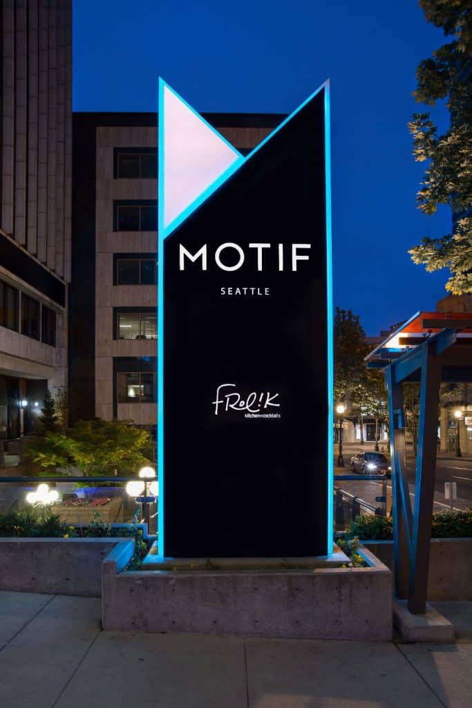 A modern, illuminated sign in an urban area reads "Motif Seattle" with "Frolik Kitchen+Cocktails" below it, set against a backdrop of buildings and trees at dusk.