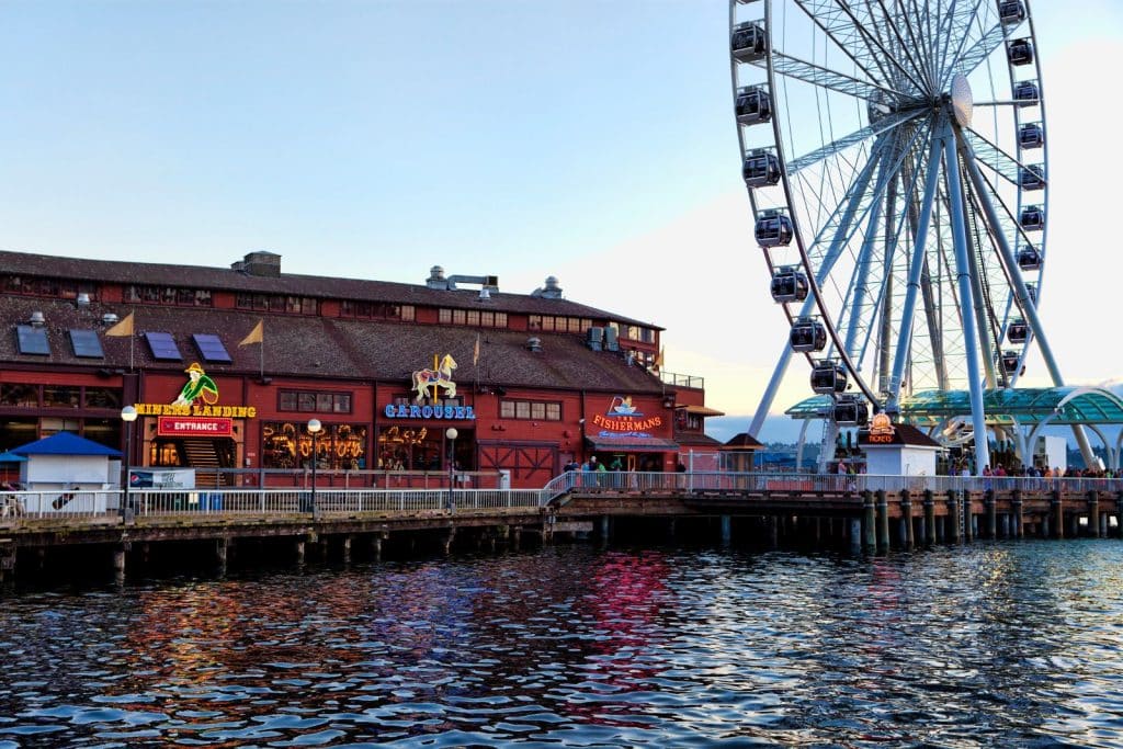 A waterfront scene with a large Ferris wheel, a building featuring a carousel, restaurants, and shops. Reflections ripple on the water below.