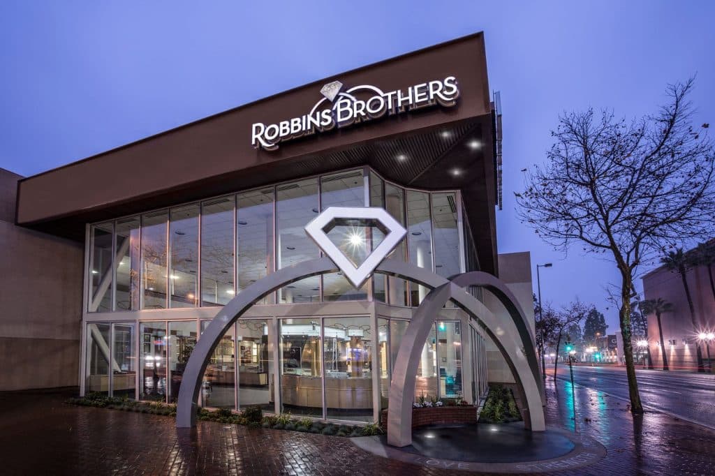 A jewelry store with a large diamond logo on the façade is illuminated at dusk, showcasing glass windows and a clear view inside. The street is wet from rain, reflecting the building's lights.