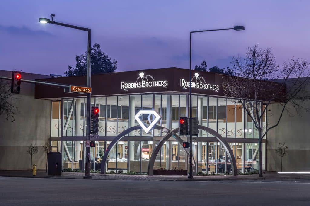 Jewelry store with a large diamond logo on the facade, located at a street corner during dusk. Traffic lights are visible in the foreground.