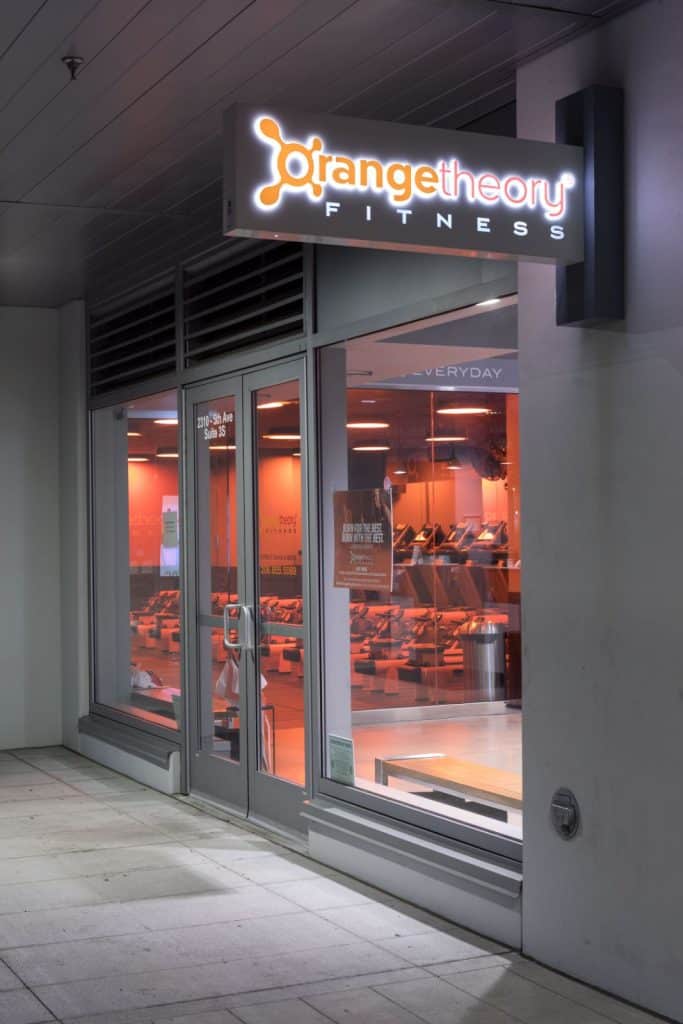 Storefront of Orangetheory Fitness with illuminated signage, glass doors, and an interior visible through the windows.