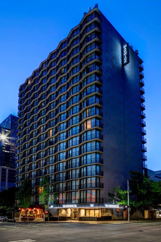 A tall, modern hotel building stands illuminated in the evening, featuring a grid-like facade with balconies. A neon sign is visible on the corner, and a street is in the foreground.