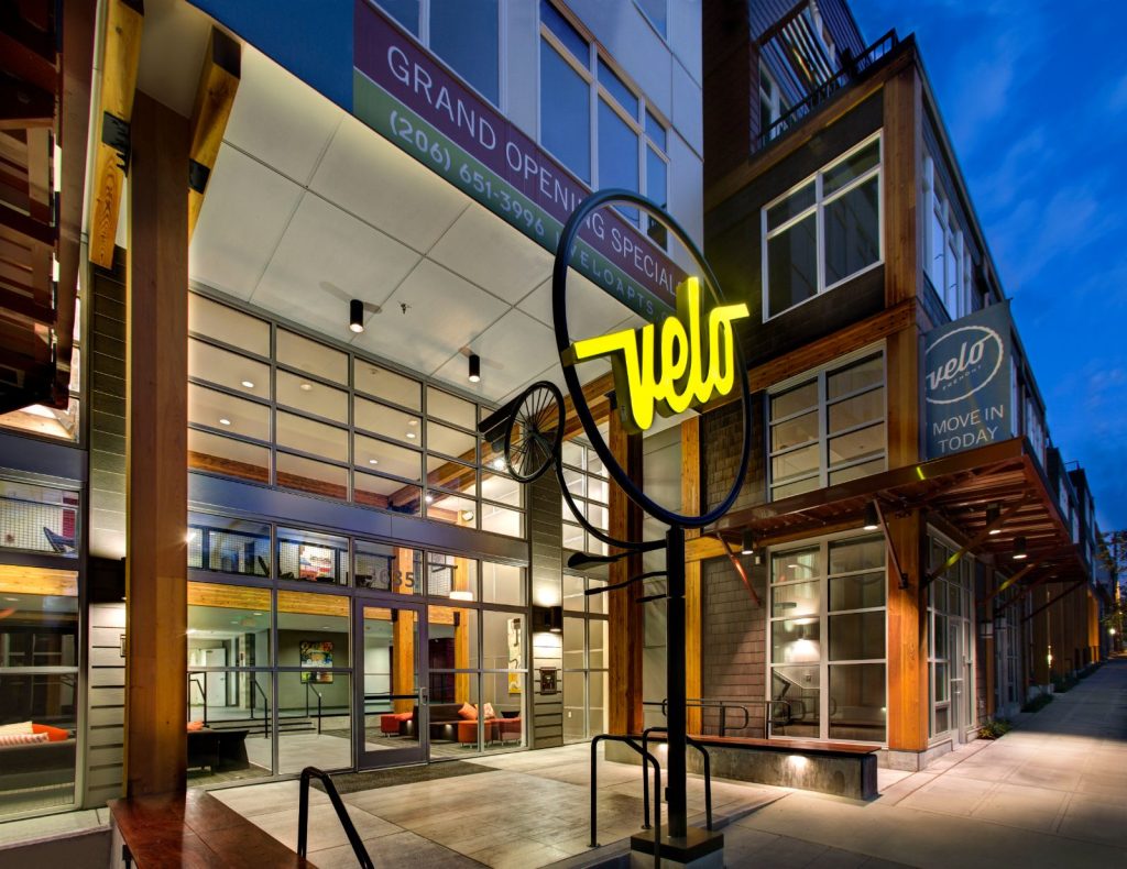 Night view of an urban apartment complex with large glass entrances and a neon sign that reads "Vuelo." A banner advertises a grand opening special.