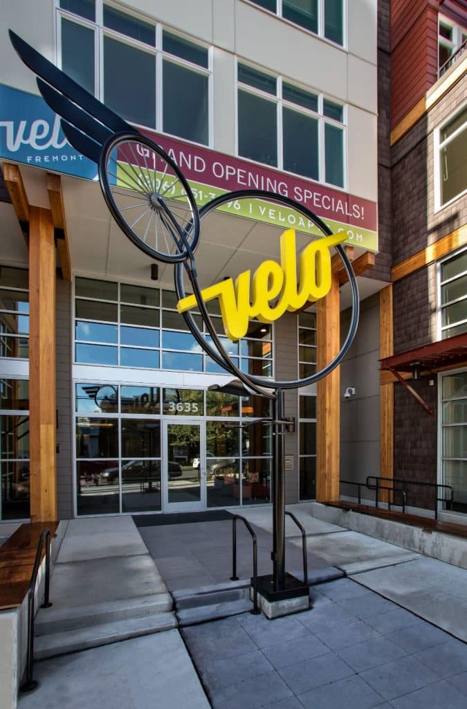 A modern building entrance featuring a large bicycle-themed sculpture with the word "velo" in yellow. Banners advertise grand opening specials.