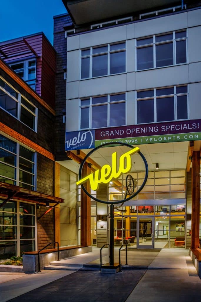 Exterior view of a modern apartment complex with a sign reading "velo" featuring a bicycle design. A banner above advertises grand opening specials.