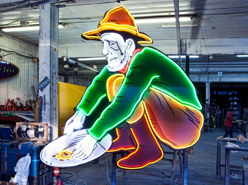 Neon sign of a man in a hat crouching and panning for gold, displayed in an industrial space.