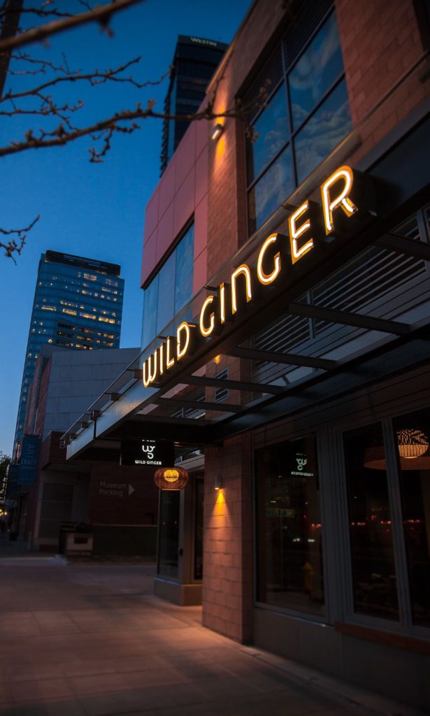 Neon-lit "Wild Ginger" sign on a modern restaurant building at dusk, with a city skyscraper backdrop.