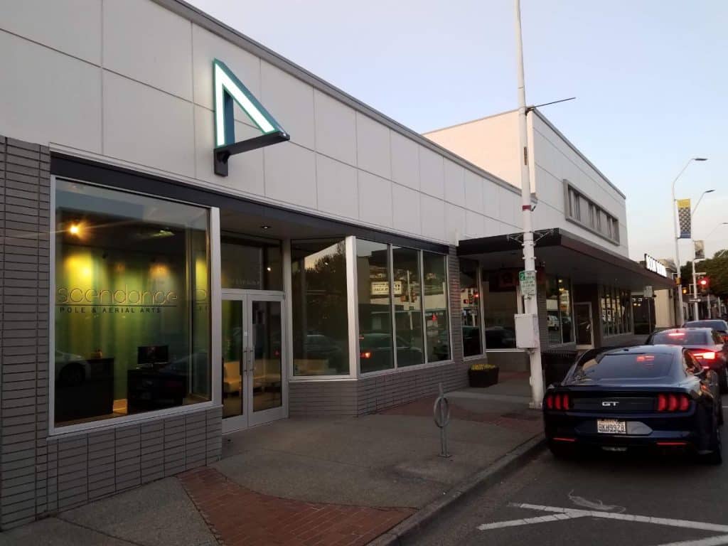 Street view of a modern commercial building with large windows and a neon triangle sign. A black car is parked on the street in front of the building.