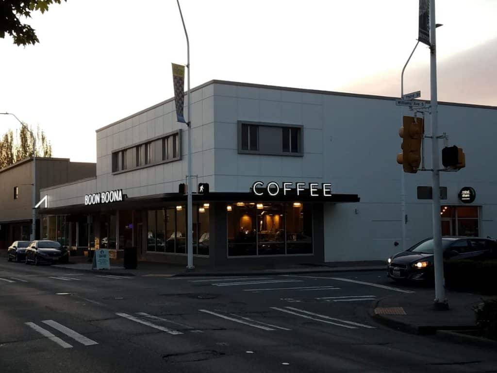 Street corner view of a building with a coffee shop and a "Boon Boona" sign, with cars parked along the road at dusk.