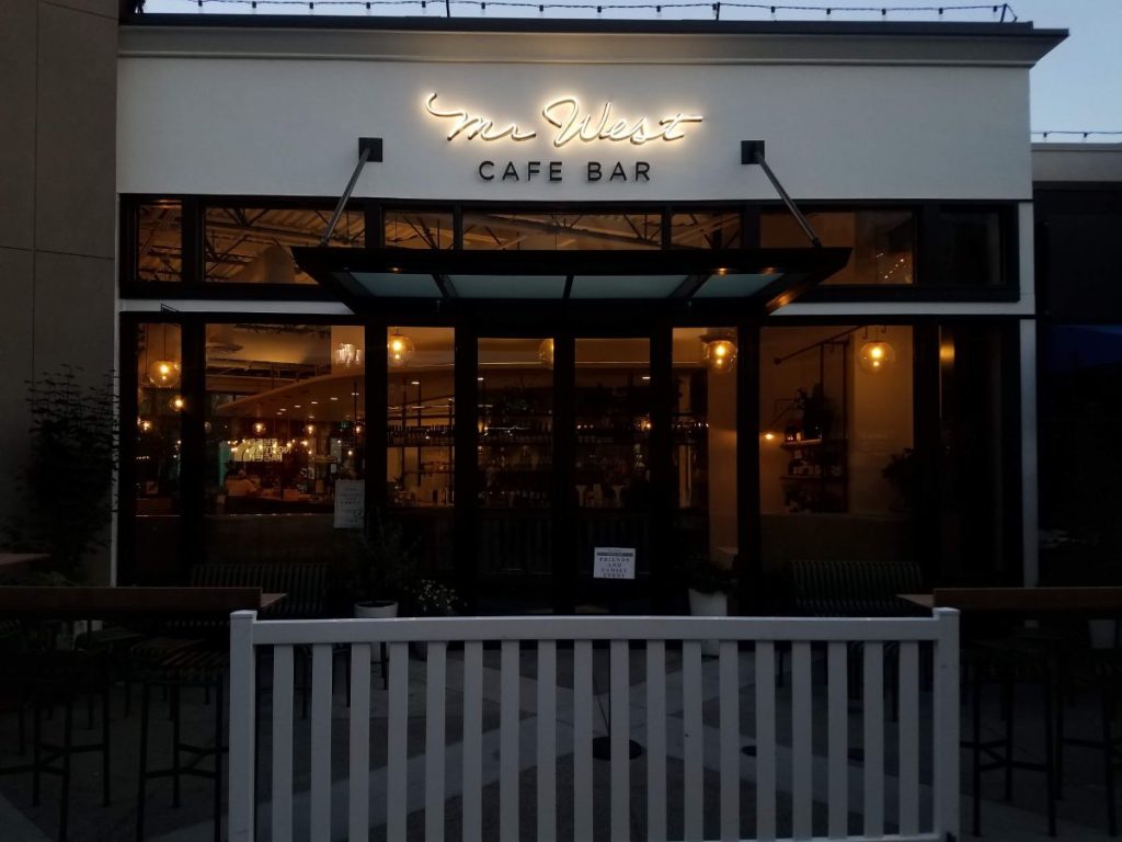 Exterior of Mr. West Cafe Bar at night with illuminated sign and a partially open glass door.