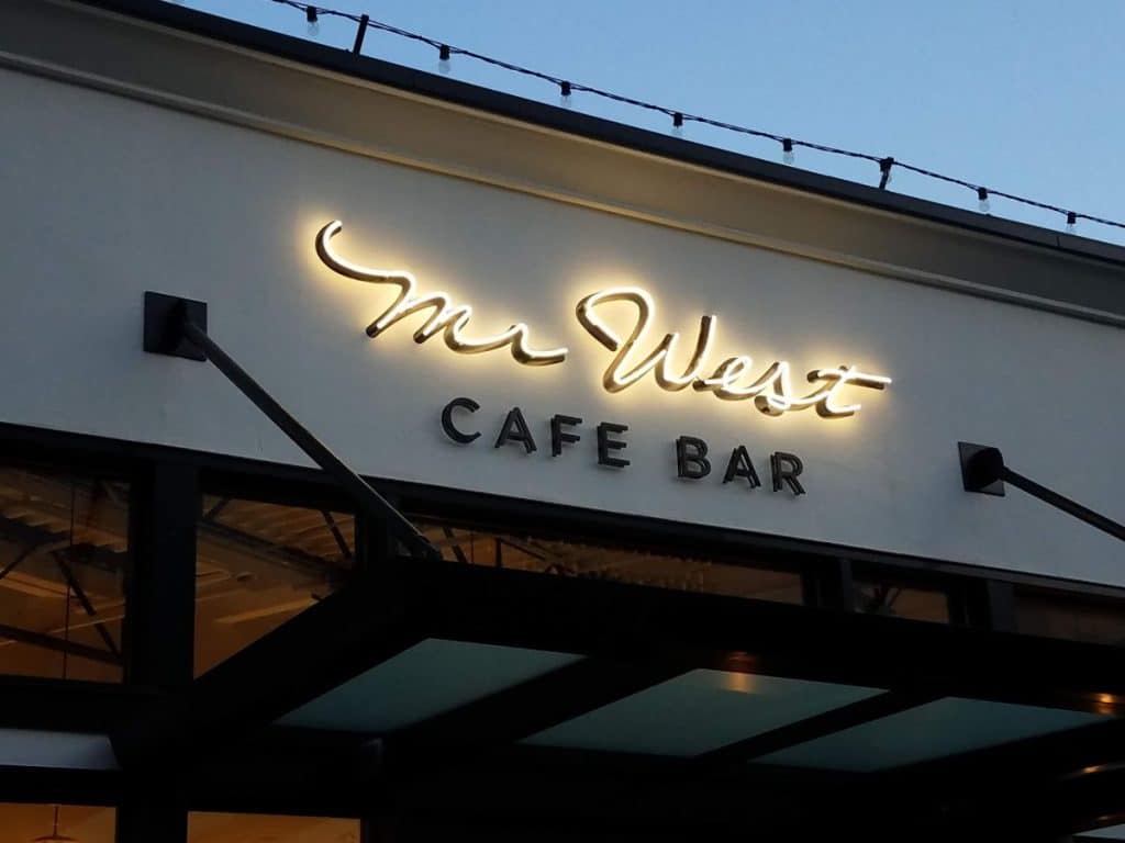 Illuminated sign for "Mr. West Cafe Bar" on a building facade, with a blue sky in the background.