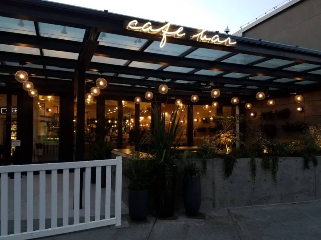 Outdoor seating area of a café with string lights and plants under a glass canopy. A sign reads "café.