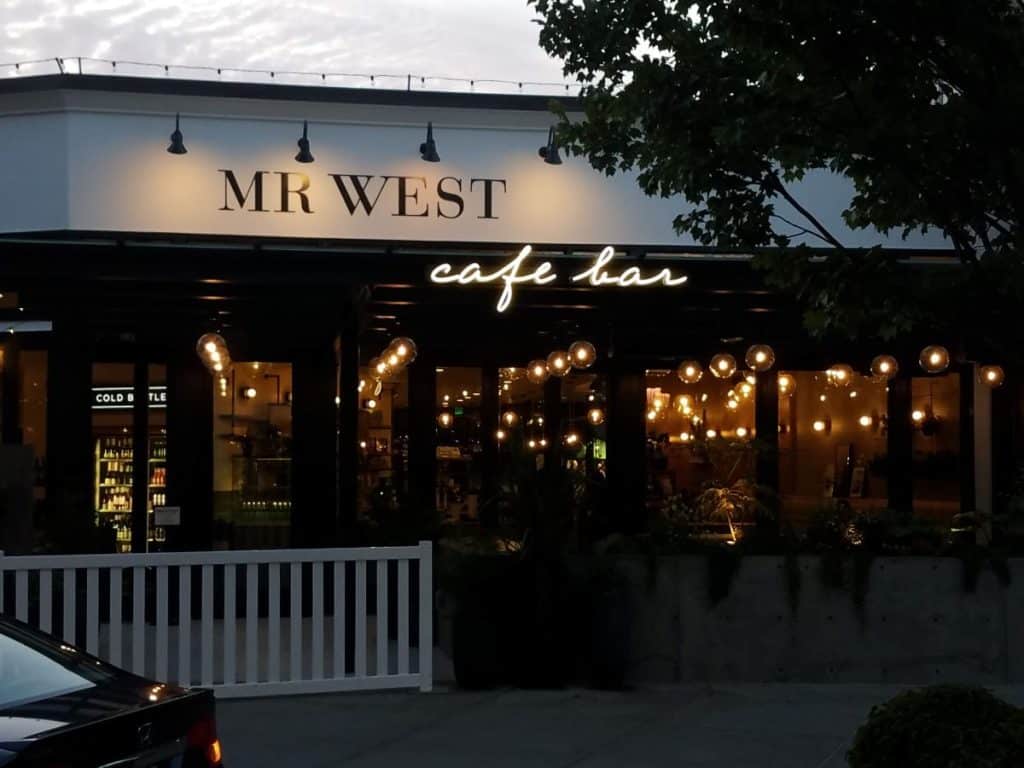 Exterior view of Mr. West cafe bar with illuminated string lights and a dark ambiance. A tree partially frames the right side.