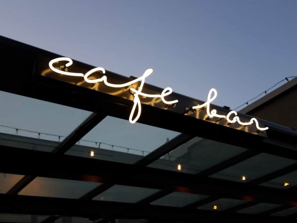 Neon sign reading "cafe bar" on a glass roof with a clear sky background.