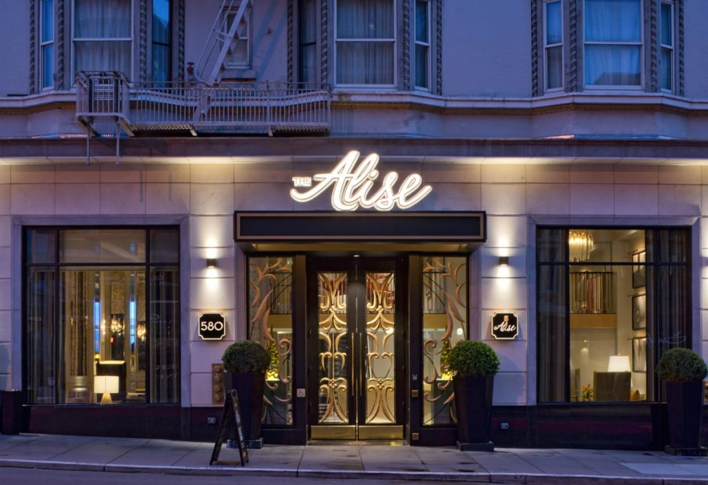 Street view of The Alise hotel entrance with illuminated sign, large windows on either side, potted plants, and a decorative glass door.