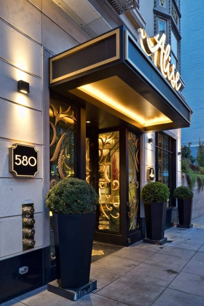 Entrance of a building with ornate glass doors, a large awning, and the number 580 displayed. Flanked by two large potted plants on either side.