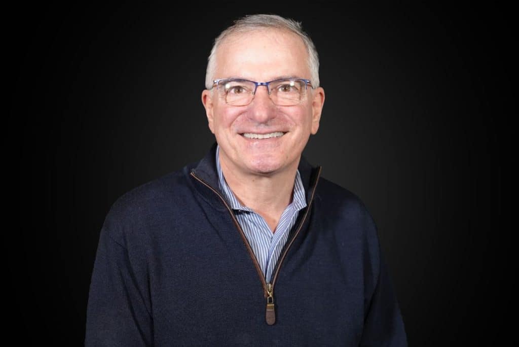 A smiling man with glasses and a blue sweater stands against a dark background.