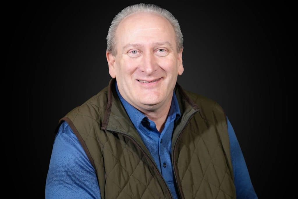 A man with short gray hair smiles at the camera, wearing a blue shirt and a brown quilted vest, set against a dark background.