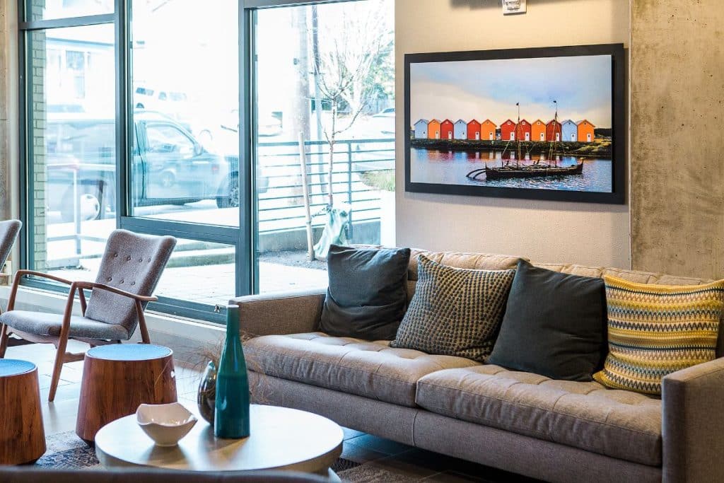 A modern living room with a gray sofa, colorful pillows, wooden stools, a round table, and a wall-mounted artwork depicting colorful buildings by the water. Large windows are in the background.