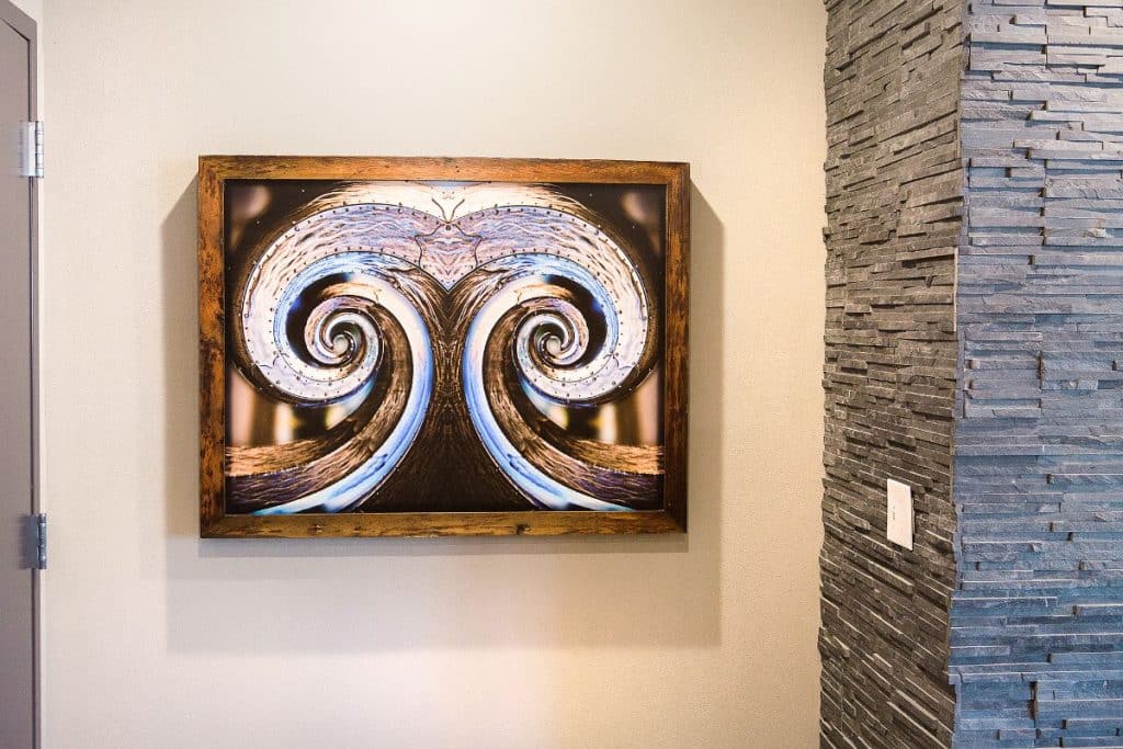 Framed artwork with swirling, mirrored abstract design on a neutral wall next to a textured stone surface.