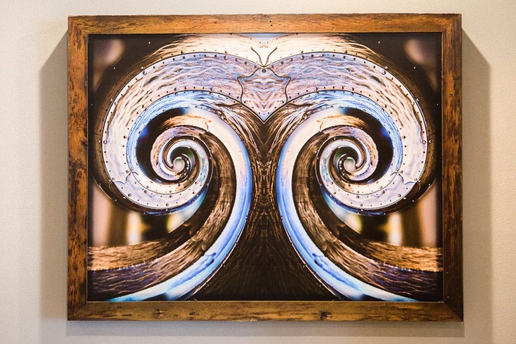 A framed abstract artwork featuring symmetrical swirling patterns with metallic and reflective textures.