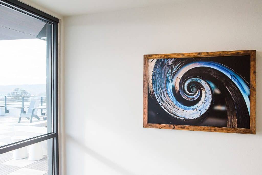 Framed spiral artwork on a white wall next to a window overlooking a balcony with a chair.