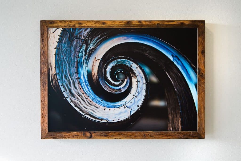 A framed abstract artwork featuring a swirling pattern with blue and brown tones, mounted on a white wall.