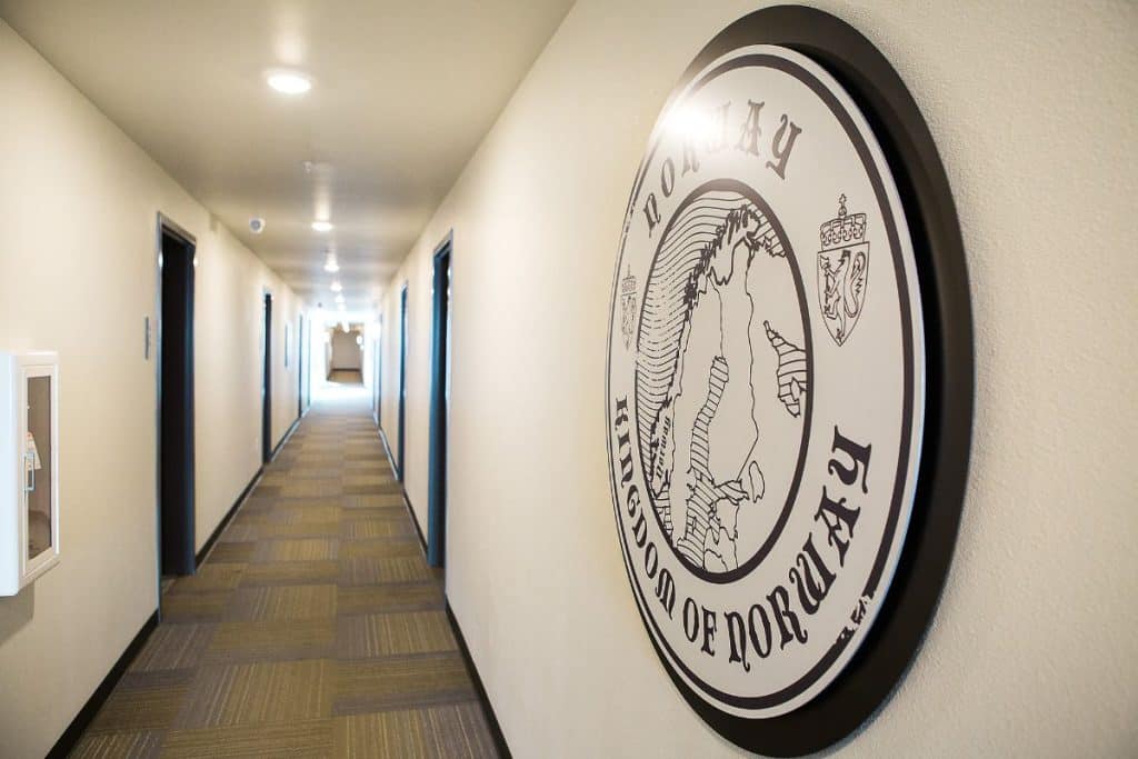 Hallway with a round sign on the wall labeled "Kingdom of Norway" and a map image. The corridor is lined with doors and has a light-toned carpet.