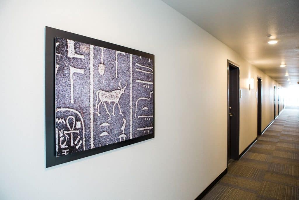 A framed artwork with ancient-style symbols, including a bull, hangs on a white wall in a well-lit hallway with patterned carpeting and several doors.