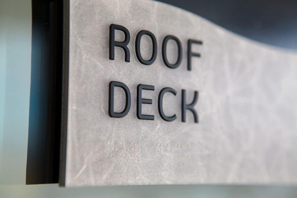 A close-up of a sign with raised black lettering that reads "ROOF DECK" on a textured, light-colored background.