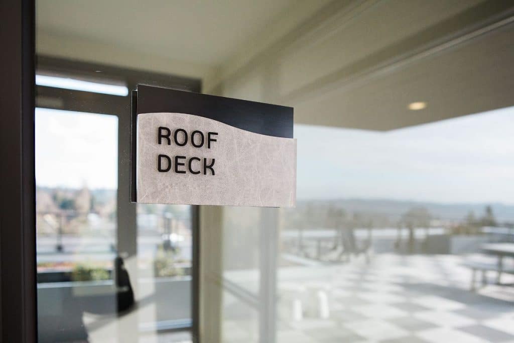 Sign reading "Roof Deck" on a glass door, with outdoor seating and a checkered floor visible through the glass.