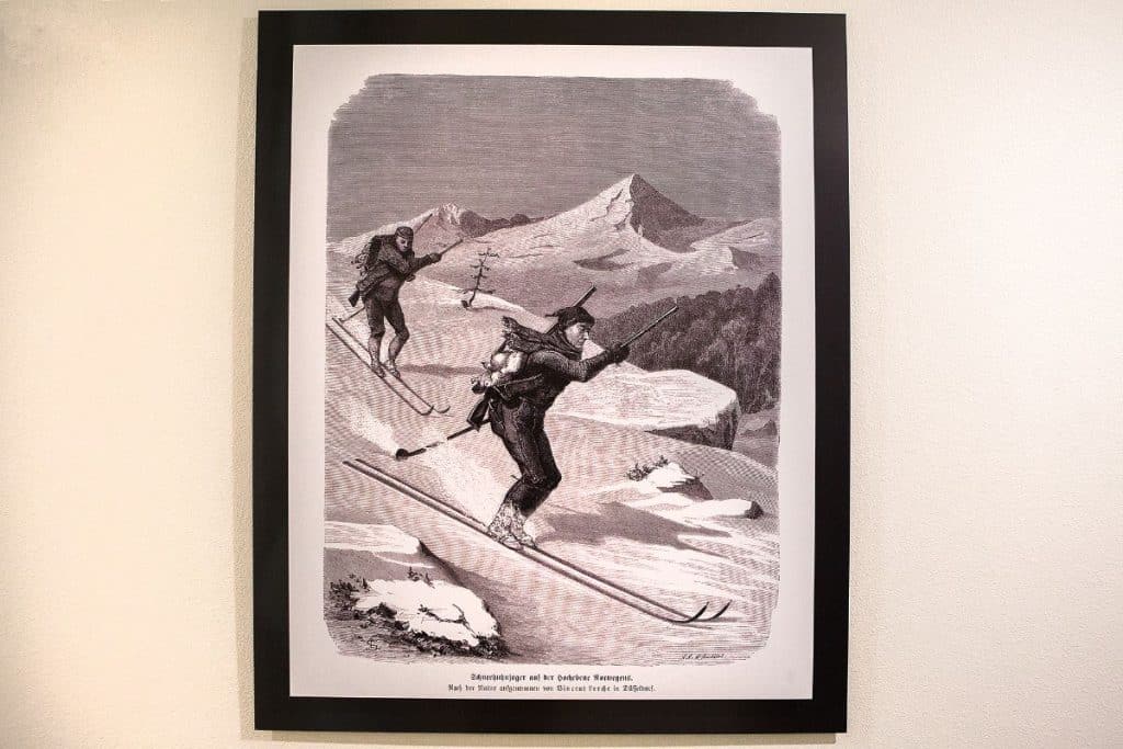 A framed illustration of two people skiing downhill with rifles on their backs, set in a mountainous snowy landscape.