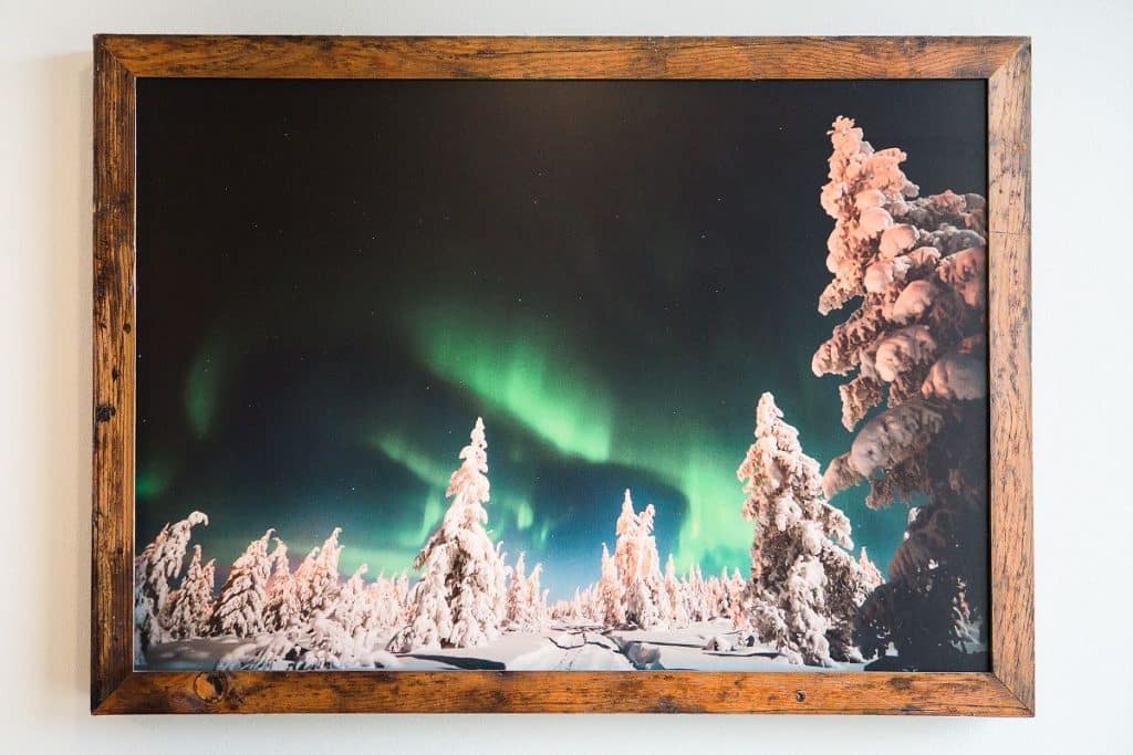 Framed photo of snow-covered trees under a vivid green aurora borealis in a dark sky.