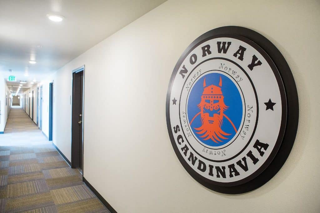 A hallway with a beige carpet and white walls featuring a circular sign on the right that reads "Norway Scandinavia" with a Viking helmet design.