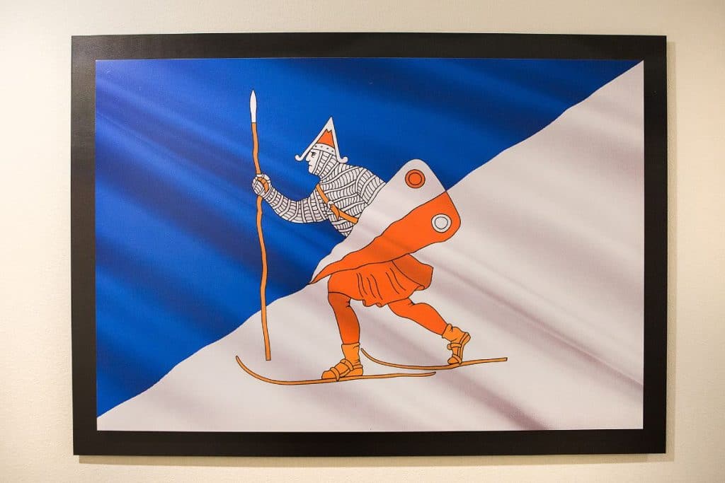 Illustration of a knight skiing downhill with a blue and white background, holding a spear and wearing armor and a red cape.